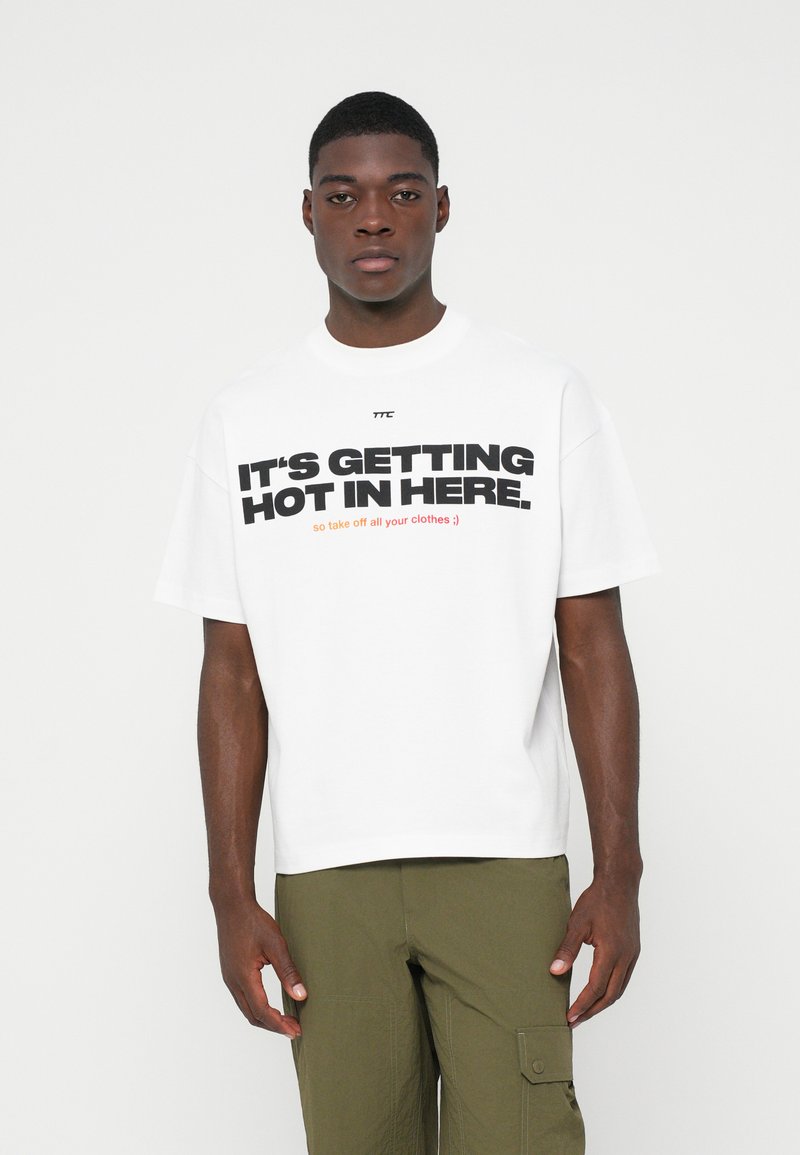 Jovem vestido com uma t-shirt branca com texto preto e laranja "ESTÁ A FICAR QUENTE AQUI" e calças cargo verde-oliva, em pé contra um fundo branco.