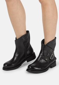 Botas de cuero negro hasta el tobillo con punta redonda, cierre de cremallera lateral y un detalle de hebilla en el exterior. Tacón bajo apilado, textura suave.