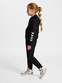 Młoda dziewczyna z rudymi włosami związanymi w kucyk, ubrana w czarną bluzę z kapturem i spodnie z białymi wzorami w szewron, oraz jasnoszare wysokie sneakersy, stojąca bokiem.