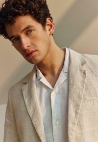 Νέος άνδρας με καστανά σγουρά μαλλιά και μπλε μάτια, φορώντας ένα μπεζ λινό σακάκι πάνω από ένα λευκό πουκάμισο με κουμπιά, κοιτάζοντας ελαφρώς προς τα κάτω.