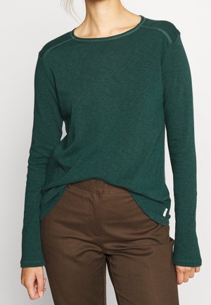 Vrouw draagt een donkergroen shirt met lange mouwen, ingestopt in hooggetailleerde bruine broek, tegen een effen achtergrond.