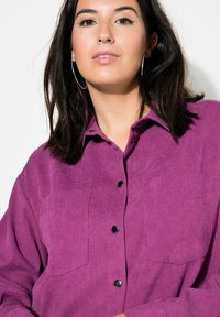 Camicia di velluto a coste viola con doppie tasche sul petto, bottoni neri e colletto classico. Texture liscia e vestibilità rilassata evidenti nel design.