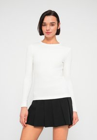 Λευκή εφαρμοστή μπλούζα με μακριά μανίκια και στρογγυλή λαιμόκοψη. Συνδυάζεται με μαύρη πλισέ φούστα, που παρουσιάζει λεία υφή και ραμμένο σχέδιο.