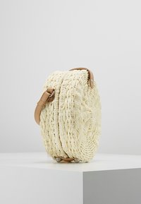 Borsa a tracolla tonda in crema intrecciata con manico in pelle beige e fibbie argentate, adagiata su una superficie bianca contro uno sfondo grigio.