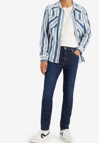 Randig blå och vit skjorta med två bröstfickor, som är kombinerad med mörkblå jeans och vita sneakers med svarta detaljer.