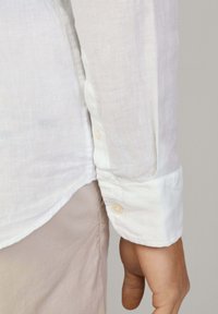 White linen shirt with a buttoned cuff. The fabric appears light and textured, with a simple design and soft finish.
