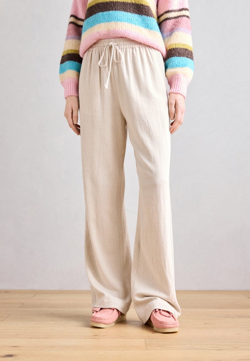 Pantaloni largi de culoare bej deschis, cu talie elastică și șnur. Textura pare moale, completată de încălțăminte roz.