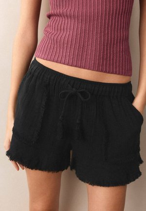 Frau trägt schwarze Shorts mit Kordelzug, ausgefransten Rändern und Vordertaschen, kombiniert mit einem ärmellosen, gerippten, mauvefarbenen Crop-Top.