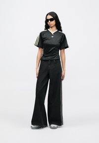 Hosszú fekete hajú nő fekete Adidas-ingben és széles szárú, oldalsávos fekete farmert viselve, fehér háttér előtt áll.