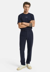 Navy katoenen T-shirt met "GANT" logo, gecombineerd met op maat gemaakte navy broeken en witte sneakers met tan accenten. Model staat met gekruiste armen.