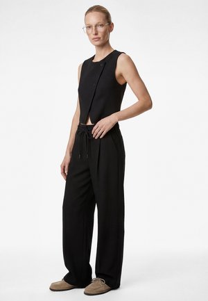 Woman wearing sleeveless black asymmetrical top, wide-leg black trousers with drawstring waist, beige lace-up shoes, and glasses, standing against white background.