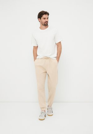 Uomo con t-shirt bianca a maniche corte, pantaloni jogger beige con coulisse e sneakers bianche, in piedi con le mani in tasca su sfondo bianco.