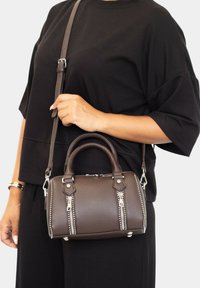 Persona con un atuendo negro sosteniendo un pequeño bolso de cuero marrón con dos asas y dos cremalleras plateadas verticales.