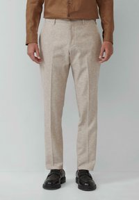 Beige maßgeschneiderte Hose aus strukturiertem Stoff, mit einem geraden Bein und vorderen Falten, kombiniert mit schwarzen Schuhen.
