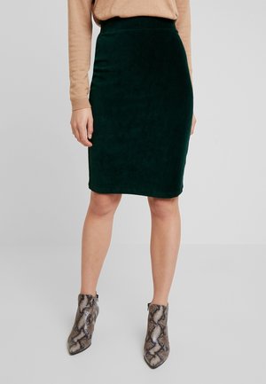 Kvinde iført en mørkegrøn knælængde nederdel, beige bluse med lange ærmer og ankelstøvler med slangeprint, stående mod en ensfarvet baggrund.