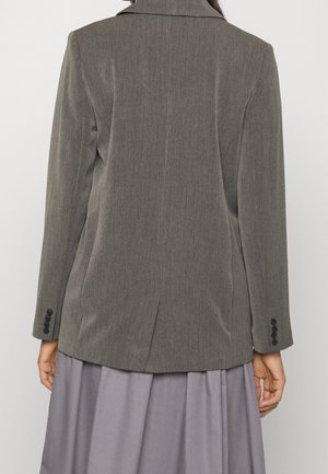 Achteraanzicht van een persoon die een nauwsluitend grijs colbert draagt met gesloten mouwen over een losse grijze rok.