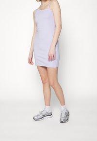 Kvinna i en lila ärmlös klänning och silverfärgade Nike-sneakers med vita strumpor, stående framför en enkel vit bakgrund.