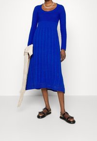 Robe tricotée bleue avec un corsage ajusté et de longues manches, présentant des motifs verticaux et une longueur mi-mollet. Portée avec des sandales noires et un sac léger.