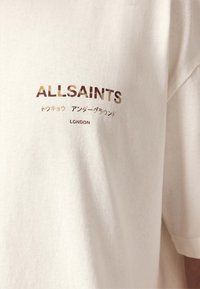 Κρεμ βαμβακερό T-shirt με κοντά μανίκια. Διαθέτει εκτυπωμένο κείμενο σε μαύρο και μεταλλικό χρυσό, περιλαμβάνοντας τις λέξεις "ALLSAINTS" και "LONDON".
