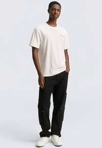 Roomwit katoenen T-shirt met korte mouwen, gecombineerd met zwarte cargobroek met zijzakken en drukknopen. Draagt witte sneakers.