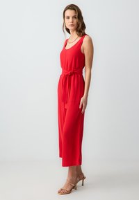 Roter ärmelloser Jumpsuit mit einem Bindeband in der Taille, entspannter Schnitt und weitem Bein. Getragen mit transparenten Riemchen-High-Heels.