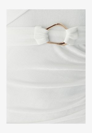 Valkoinen kangasmekko, jossa on solmittu vyö ja kultainen metallikoriste. Materiaali on pehmeää, teksturoitua ja siinä on hienovaraiset laskokset.