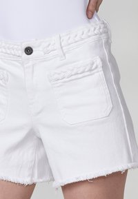 Pantalones cortos de mezclilla blanca con detalle de cinturilla trenzada, costuras de contraste, dobladillo deshilachado y dos bolsillos frontales. Elaborados con tela de algodón texturizada.