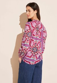 Long-sleeve blouse in vibrant pink, purple, and red geometric pattern, with a round neckline and gathered back detail, paired with wide-leg denim.