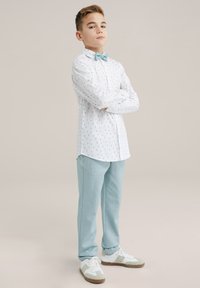 Camicia bianca con dettagli a motivi verdi, abbinata a pantaloni della tuta azzurri chiari e sneakers bianche con dettagli grigi e marroni.