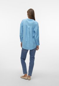Camicia blu a maniche lunghe con una vestibilità rilassata, polsini abbottonati e pieghe sul retro, abbinata a jeans blu chiaro e ballerine beige.