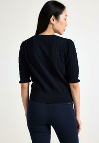Maglione navy in maglia con texture a coste e maniche corte a palloncino. Il maglione presenta bordi ondulati e una vita aderente per una vestibilità sagomata.