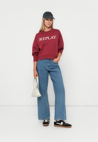 Sweat-shirt bourgogne avec logo "REPLAY", jean large bleu clair, baskets noires avec accents blancs, et casquette grise, portant un petit sac blanc.
