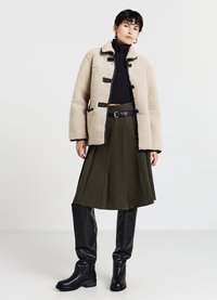 Giacca in shearling beige con bottoni neri, dolcevita a coste nere, gonna plissettata oliva e stivali neri al ginocchio, creando un look a strati.