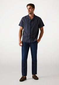 Camisa de manga corta azul marino con rayas blancas verticales, combinada con pantalones vaqueros oscuros y mocasines marrones; tejido suave y corte relajado.