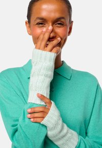 Femme souriante, portant des gants tricotés sans doigts de couleur bleu clair et un pull teal avec un col, sur fond blanc.