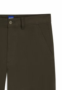 Close-up van donker olijfgroene broek met zijzakken, ceintuurlussen, metalen knoop en zichtbaar blauw HUGO-merklabel.