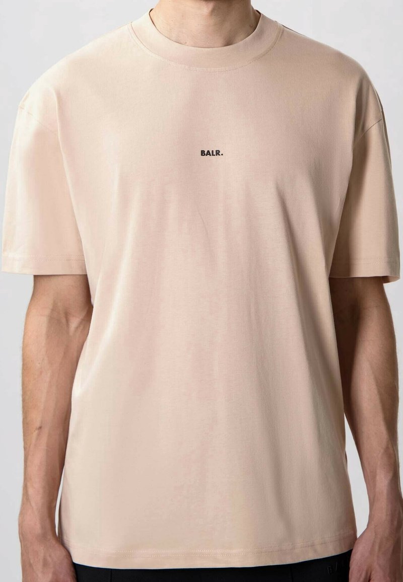 T-shirt beige à manches courtes avec petit logo noir "BALR." centré sur la poitrine, porté par une personne avec des bras visibles.