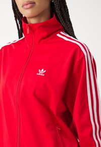 Giacca sportiva rossa con zip, realizzata in materiale sintetico liscia, con strisce bianche sulle maniche e un piccolo logo bianco sul petto.