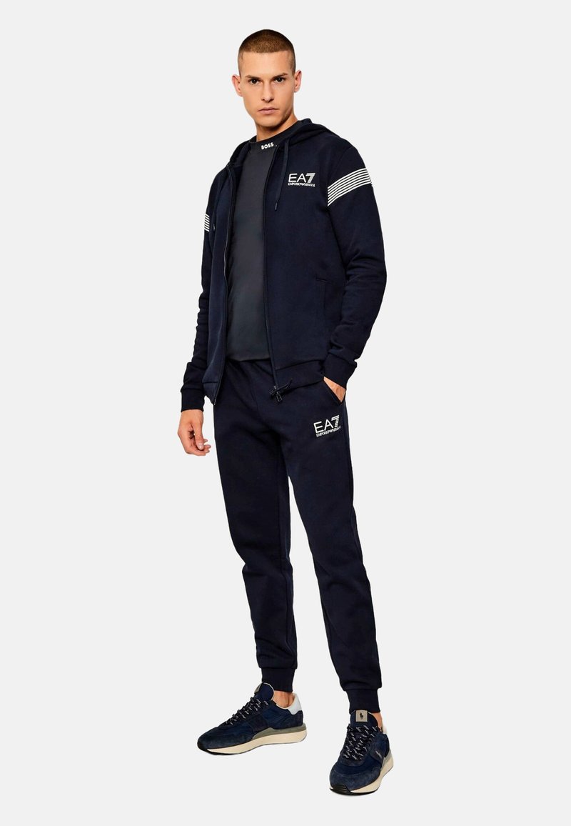 Felpa con cappuccio blu scuro con zip e pantaloni jogger abbinati in tessuto morbido, caratterizzati da strisce bianche sulle maniche e loghi sul petto e sui pantaloni.