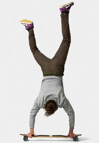 Een skateboard met een houten deck en zwarte grip tape, ondersteboven vastgehouden door een persoon in een grijze sweatshirt en een bruine broek, die een handstand uitvoert.