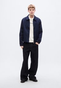 Ναυτικό μπουφάν με φερμουάρ και γιακά-ανοιχτό, φορεμένο πάνω από λευκό μπλουζάκι με μαύρο κείμενο, σε συνδυασμό με φαρδιά σκούρα τζιν και μαύρα παπούτσια.