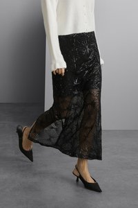 Jupe en dentelle noire avec un motif géométrique, en tissu transparent, longueur genoux ; associée à des talons slingback noirs à bout pointu et à un pull blanc.