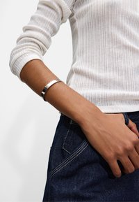 Srebrna bransoletka na rękę o gładkim wykończeniu i małych wypukłych kropkach, noszona na nadgarstku, obok fakturowanej, białej bluzki w prążki oraz ciemnoniebieskich dżinsów.