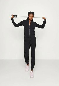 Melnas rāvējslēdzēja jakas un atbilstošas jogger bikses, abas izgatavotas no mīksta auduma. Modelis tur fitness aksesuāru, ģērbies pasteļrozā sporta apavos.