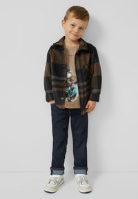 Junger Junge, der eine braune karierten Jacke, ein T-Shirt mit Scooter-Design, dunkle Jeans und weiße Sneakers trägt und vor einem schlichten Hintergrund steht.