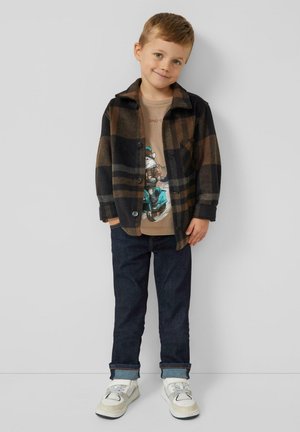 Jongentje in een bruine geruite jas, graphic t-shirt met een scooterontwerp, donkere jeans en witte sneakers, staand tegen een effen achtergrond.