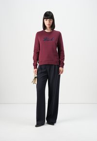 Donna in piedi che indossa un felpa bordeaux con la scritta "Karl", pantaloni neri a gamba larga, scarpe nere e tiene in mano una piccola borsa beige.