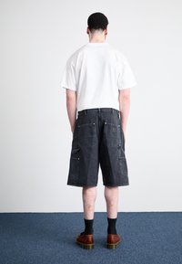 Black denim cargo shorts with multiple back pockets, complementing a white T-shirt. Worn with black socks and brown shoes with a chunky sole.