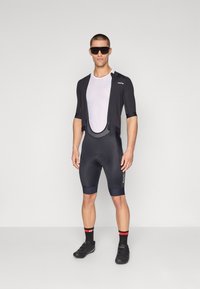 Herre sykling antrekk med en svart kortermet trøye over et hvitt mesh-bunnlag og svarte bib-shorts med reflekterende detaljer.