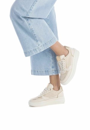 Zapatillas beige con textura trenzada, suela blanca y puntera redonda. Combinadas con pantalones de mezclilla de pierna ancha en azul claro. Diseño simple y limpio.